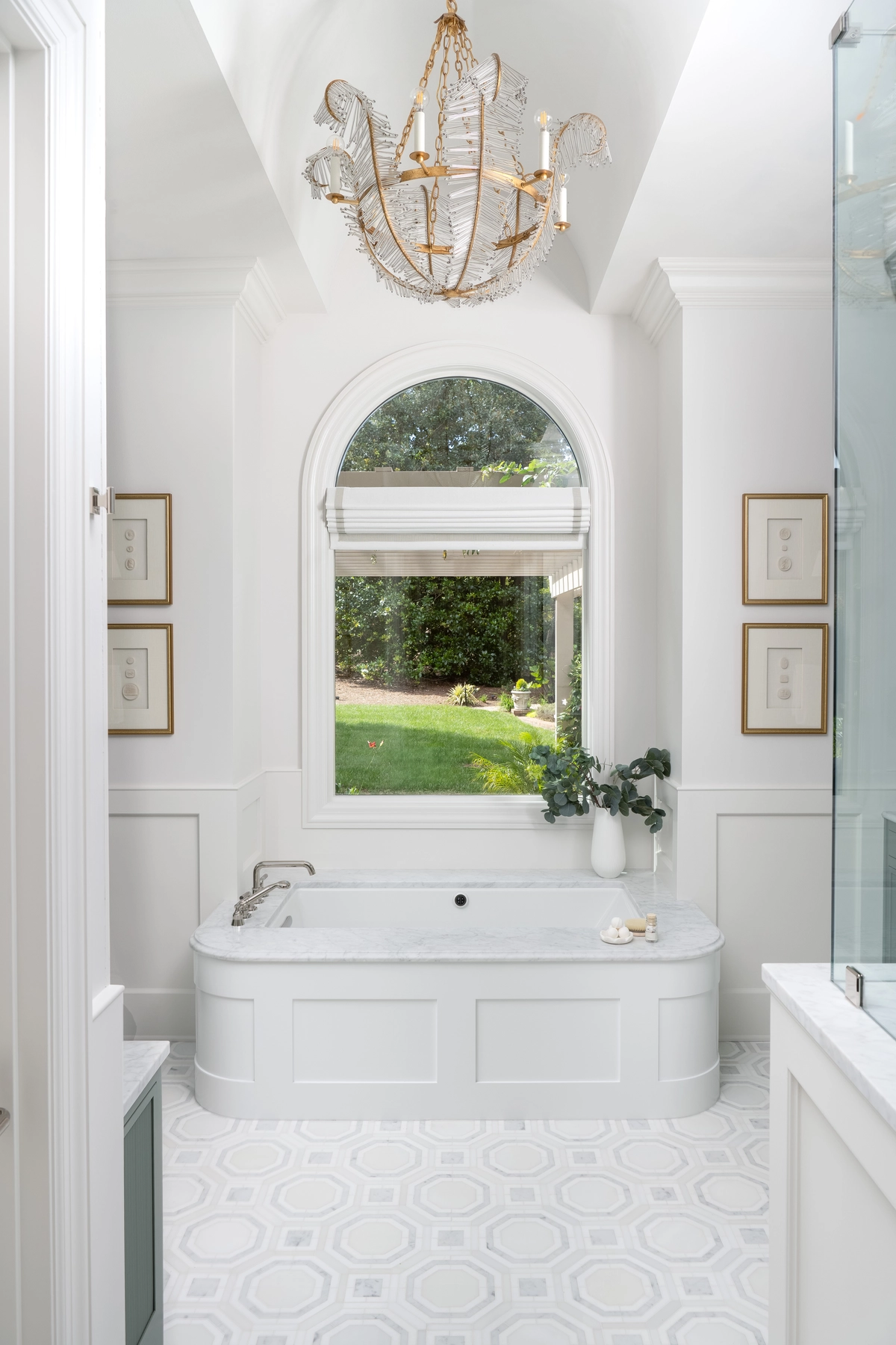 Luxurious white bathroom with a paneled soaking tub beneath an arched window, ornate gold crystal chandelier, and geometric tile flooring
