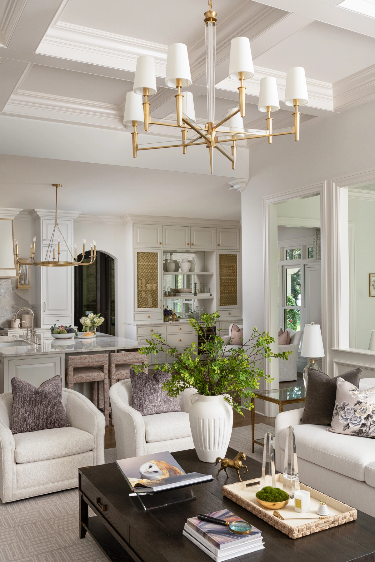 Elegant living room with a coffered ceiling, brass multi-arm chandelier, cream upholstered armchairs, dark wood coffee table, and open sightlines into the white kitchen