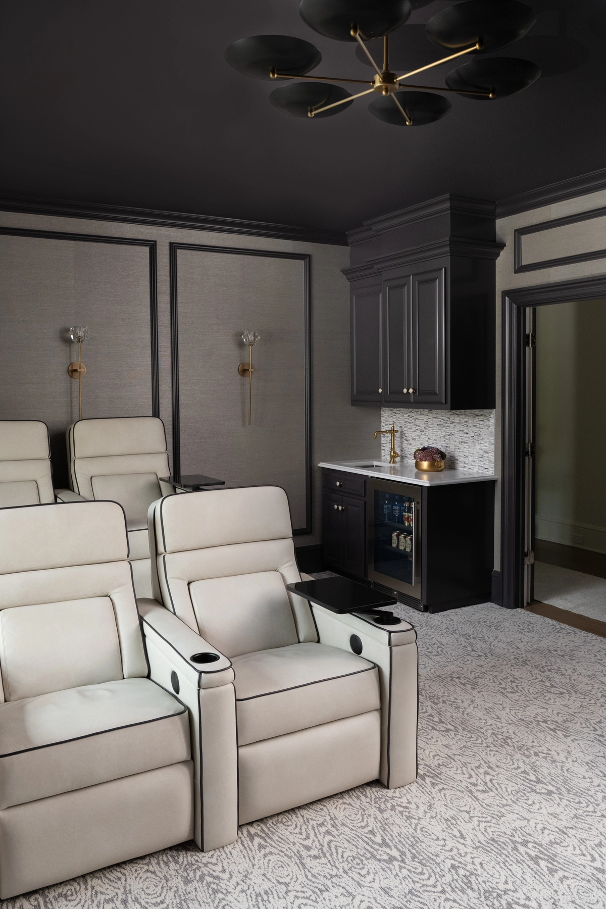 Home theater room with dark charcoal walls, cream leather reclining seats, a built-in beverage bar, patterned carpet, and a modern brass ceiling fan