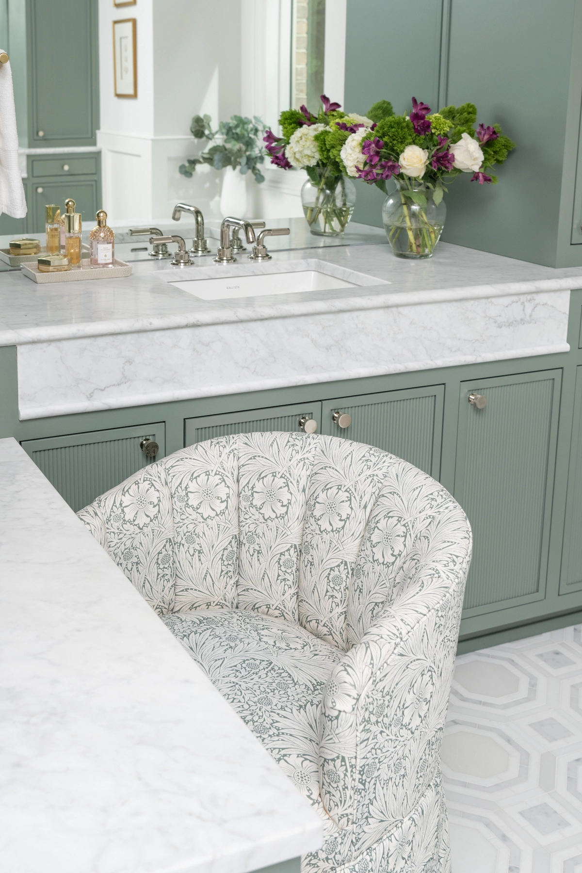 Close-up of a sage green bathroom vanity with white marble countertop, polished nickel faucet, floral upholstered vanity chair, and fresh purple and white flowers