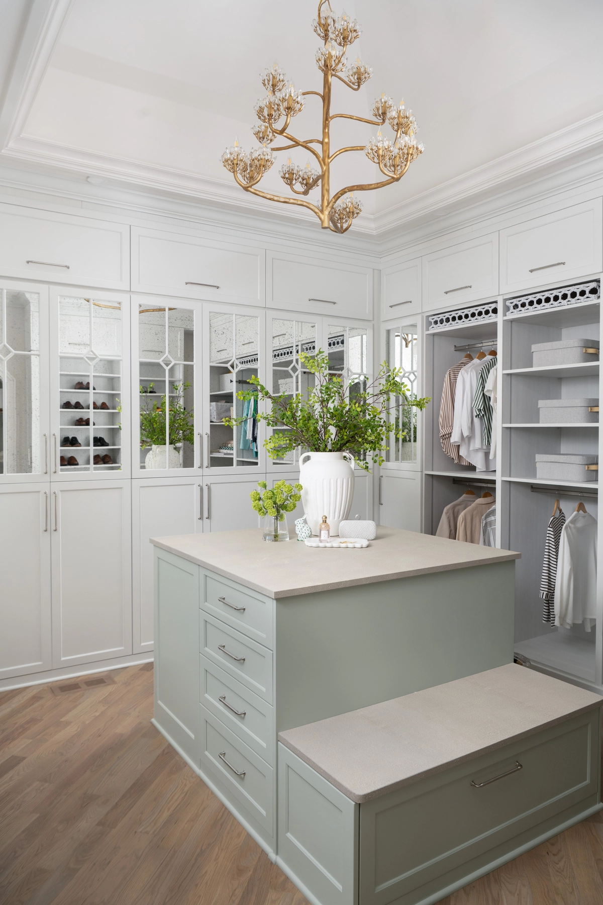 Spacious walk-in closet with a sage green center island, white mirrored glass-front cabinetry, gold floral chandelier, and warm wood flooring