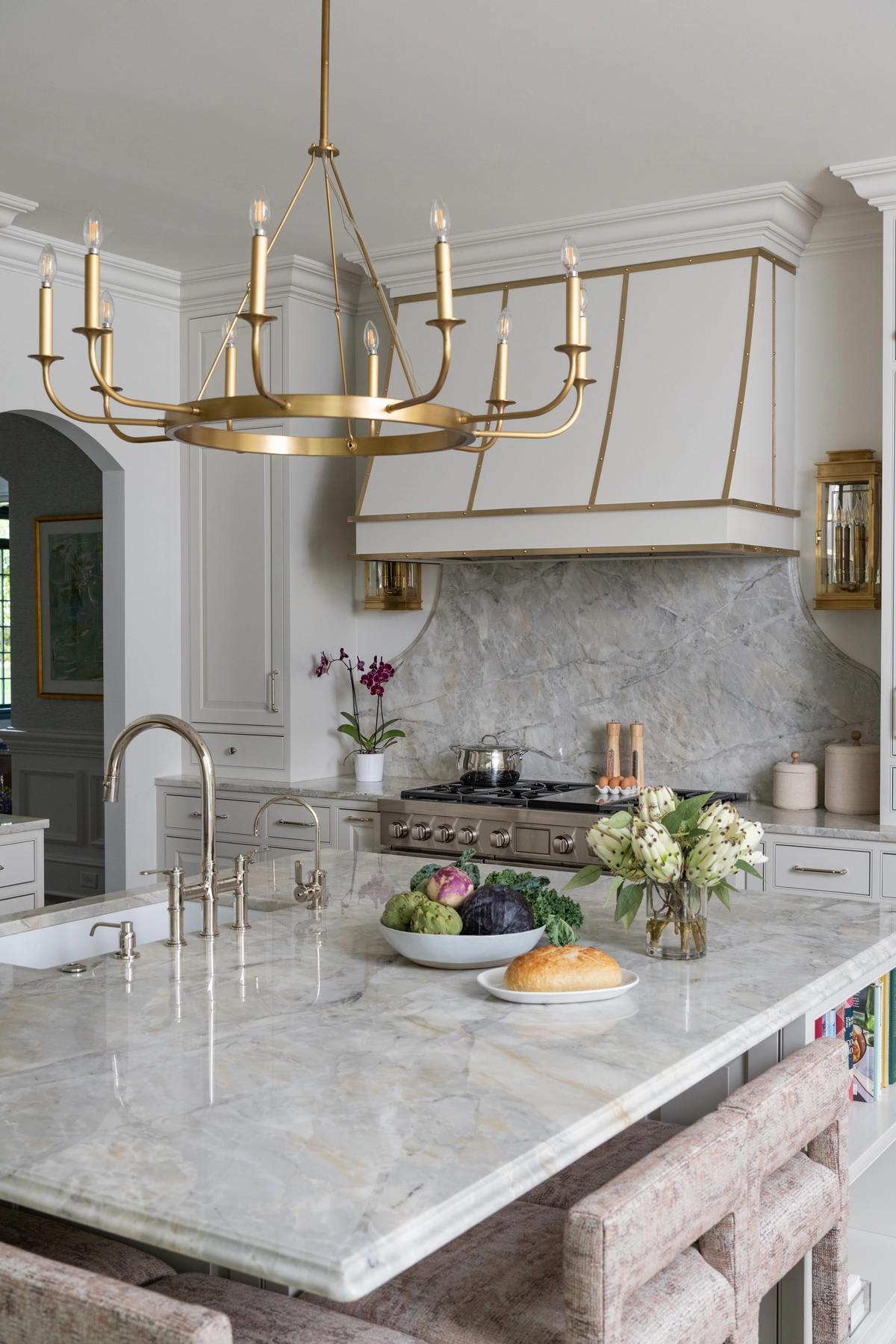 Gourmet kitchen featuring a marble-topped island with brass bridge faucet, a large brass candelabra chandelier, white and brass range hood, and marble backsplash