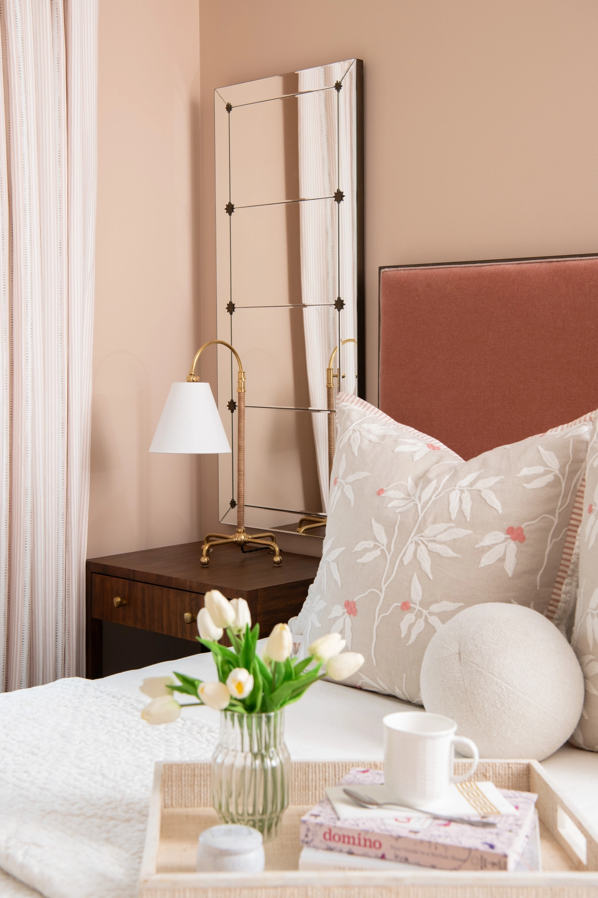 Warm bedroom vignette with a peach velvet headboard, dark wood nightstand, brass swing-arm lamp, and a glass vase of white tulips on a breakfast tray