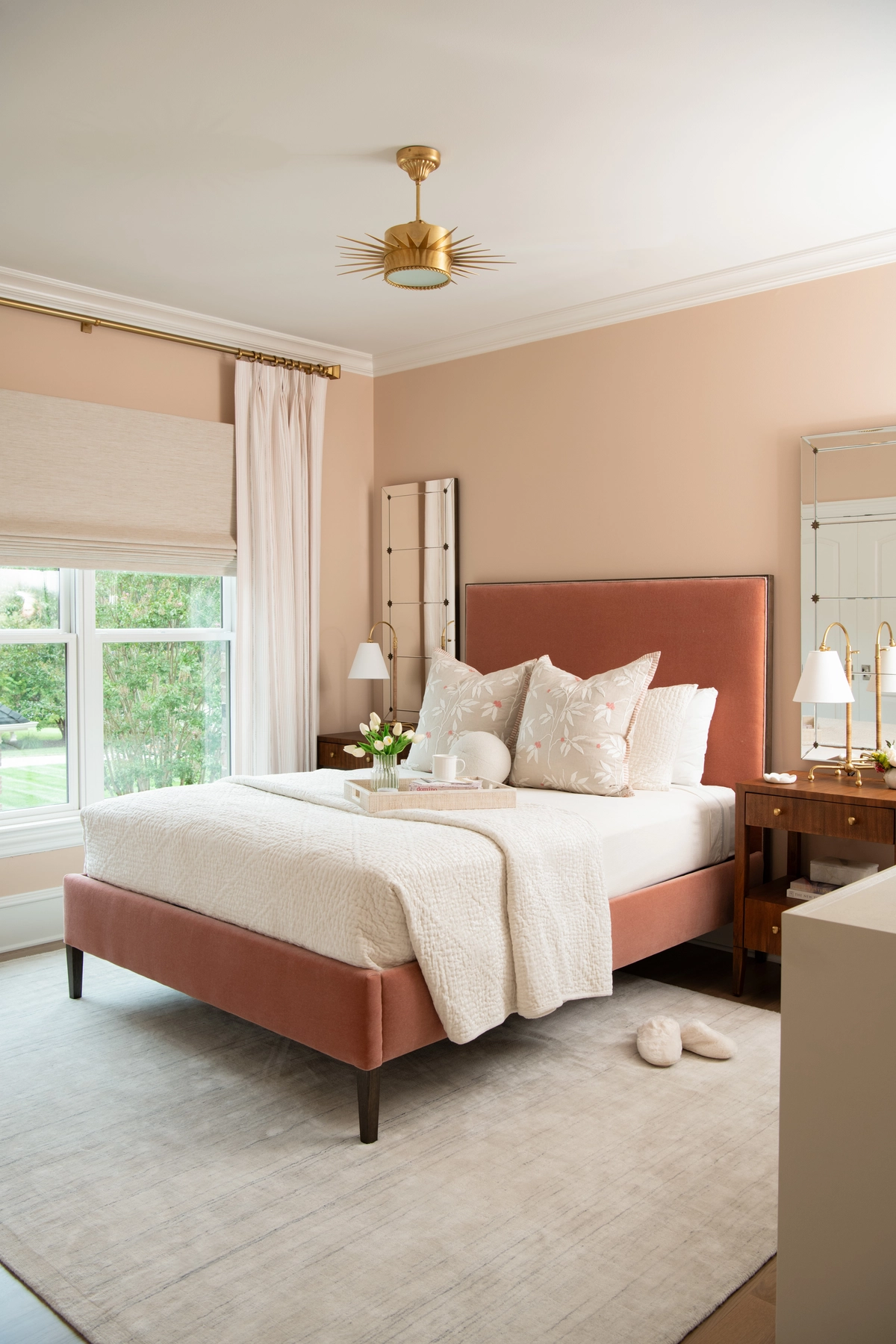 Inviting bedroom with soft peach walls, a rust-toned velvet upholstered bed, white textured bedding, floral accent pillows, and a gold sunburst flush-mount light