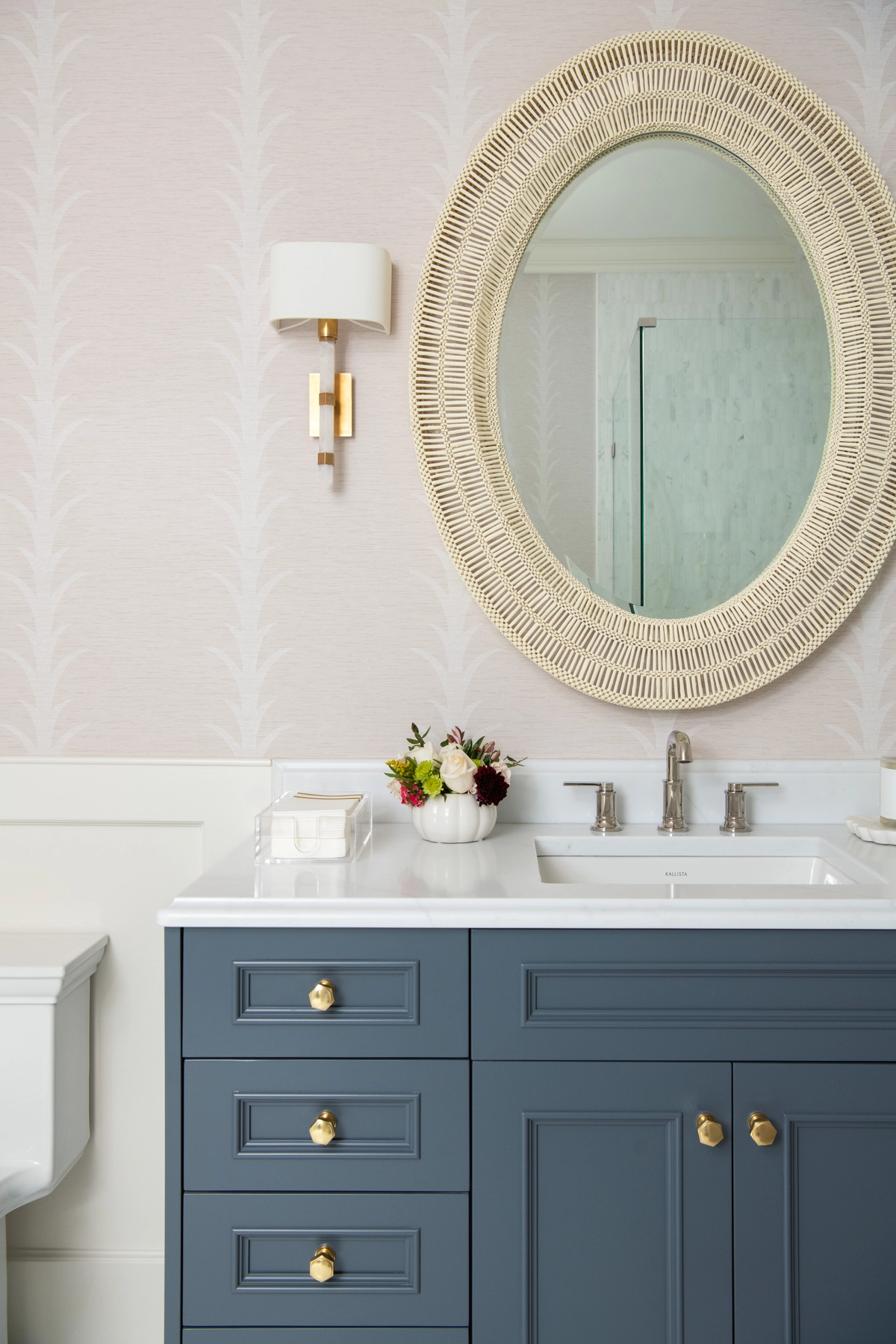 Feminine bathroom with blush pink Art Deco-inspired wallpaper, a slate blue vanity with gold knobs, white quartz countertop, and an oval woven mirror