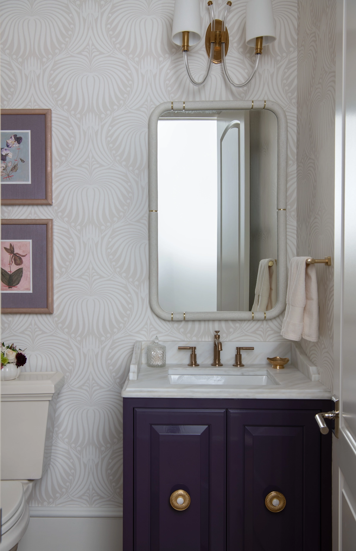 Petite powder room with a deep purple vanity, white marble top, brass hardware, subtle fan-patterned wallpaper, and framed botanical prints