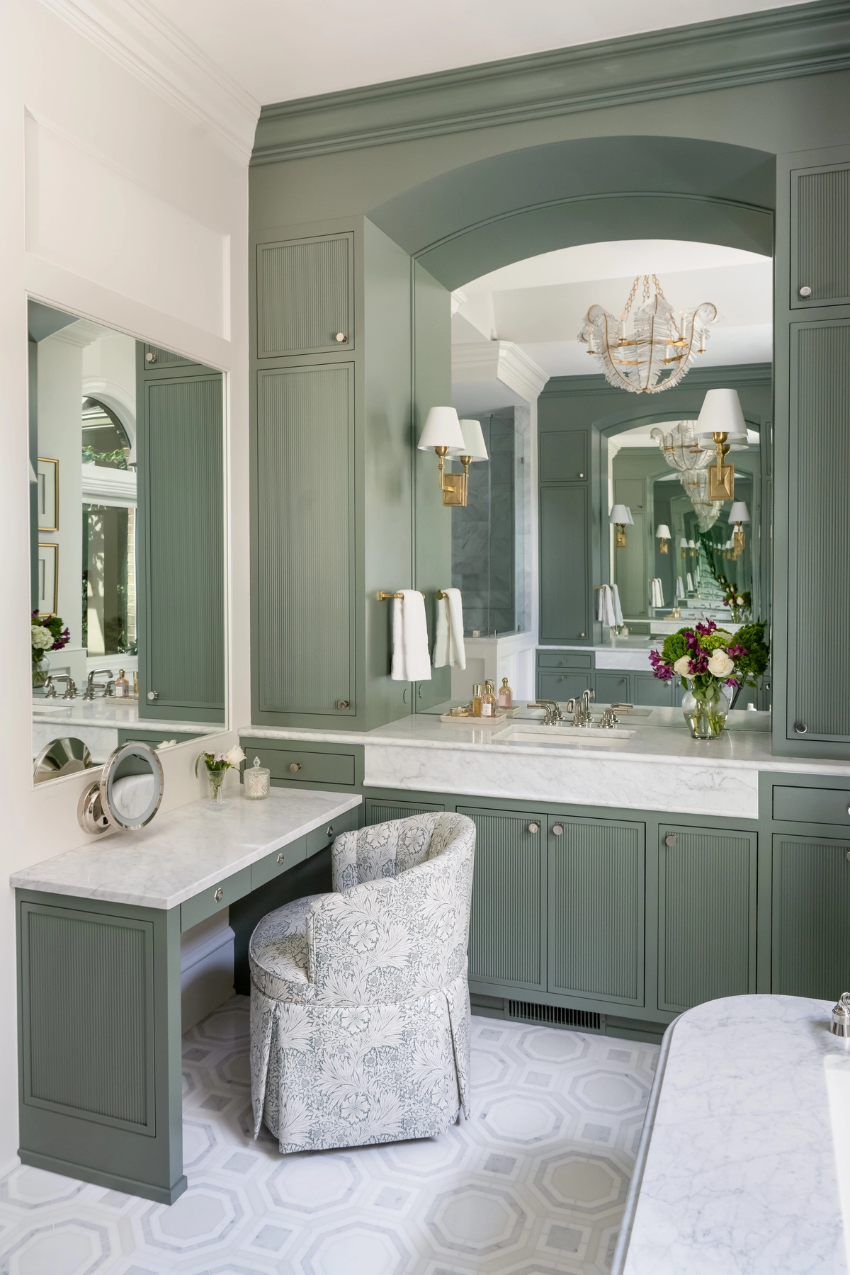 Primary bathroom with floor-to-ceiling sage green cabinetry, an arched mirror alcove, marble countertops, a built-in vanity desk, brass sconces, and geometric tile flooring