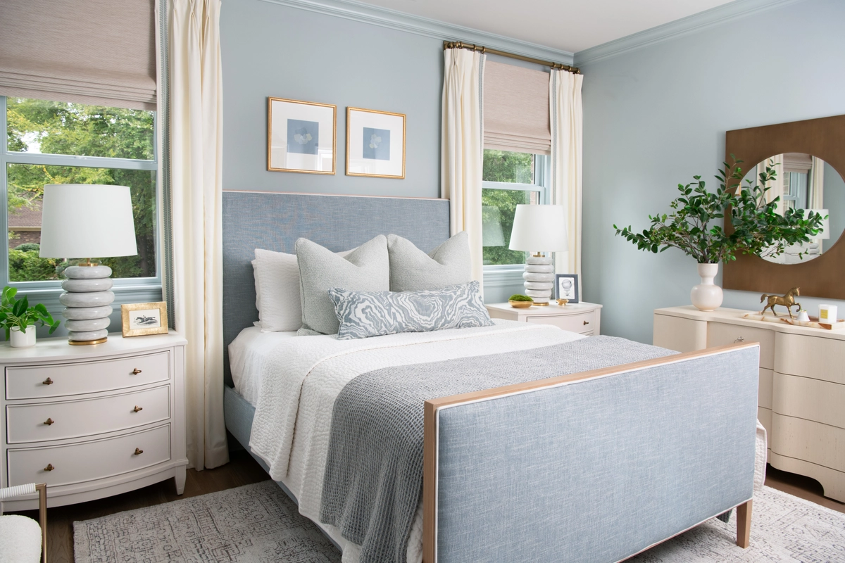 Serene blue bedroom with a light blue upholstered bed, matching blue walls, white curved nightstands with stacked ceramic lamps, and ivory curtains