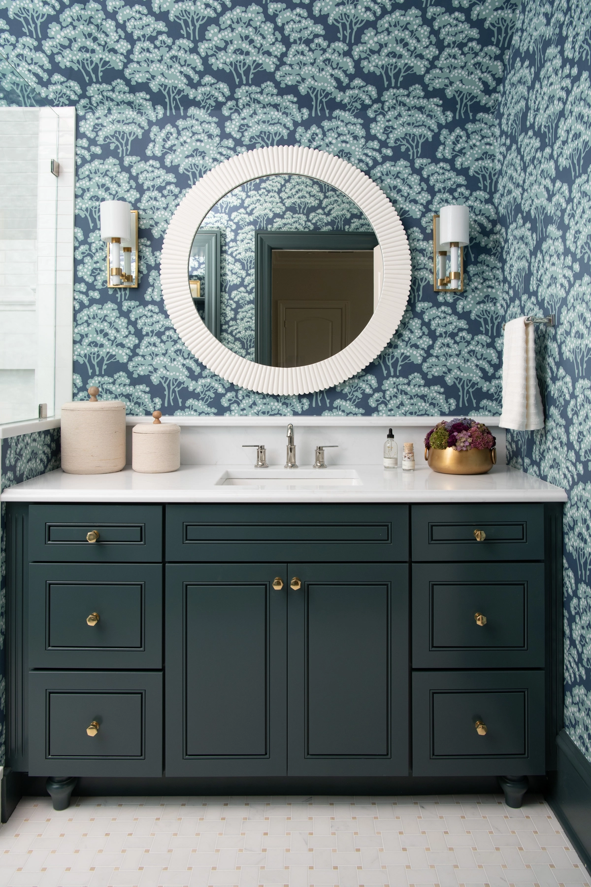 Ensuite bathroom vanity with dark teal cabinetry, brass knobs, a round white-framed mirror, teal tree-patterned wallpaper, and brass and glass wall sconces