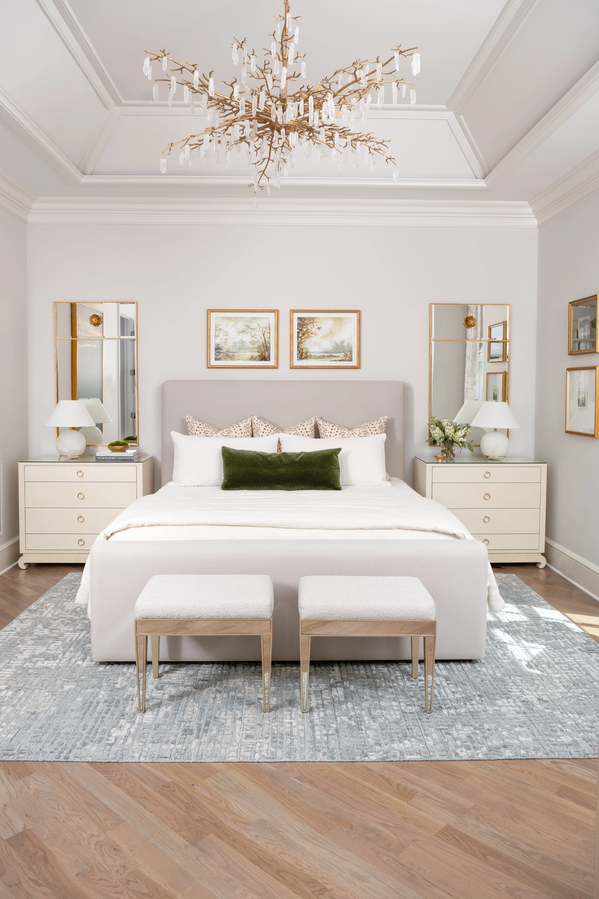 Symmetrical primary bedroom with a light gray upholstered bed, cream nightstands, tall gold-framed mirrors, a gold branch chandelier, and a soft blue area rug