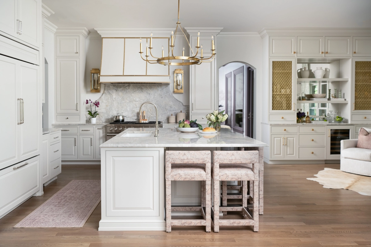 Bright white kitchen with a large center island, marble countertops, upholstered bar stools, a brass and white candelabra chandelier, and cane-front cabinet inserts