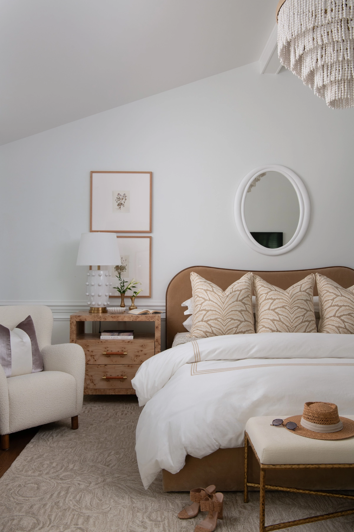 Primary bedroom viewed from the side showing a camel upholstered bed with botanical pillows, a burl wood nightstand with a white dotted ceramic lamp, and a cream bouclé armchair