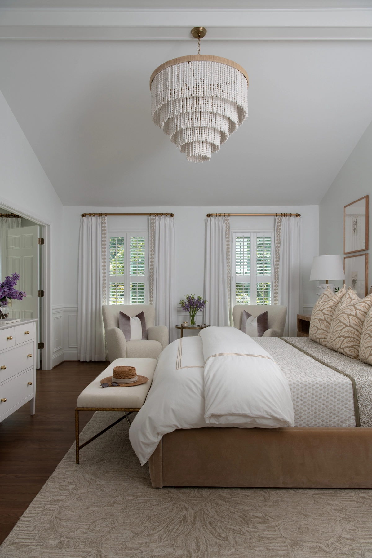 Angled view of the primary bedroom highlighting the tiered beaded chandelier on the vaulted ceiling, plantation shutter windows with white drapery, and cream armchairs in the sitting area