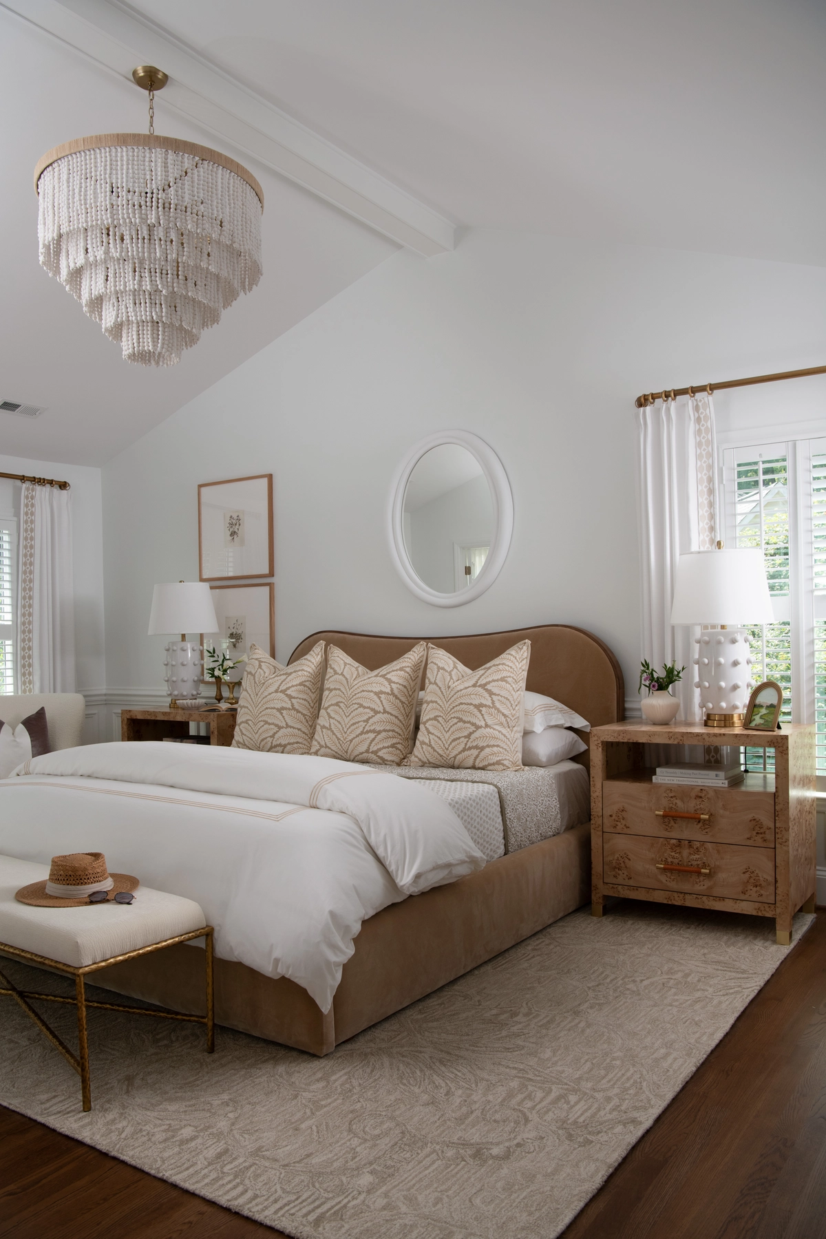 Front-facing view of the primary bedroom with a camel upholstered bed, burl wood nightstands, white dotted ceramic lamps, a white oval mirror, botanical leaf pillows, and a tiered beaded chandelier on the vaulted ceiling