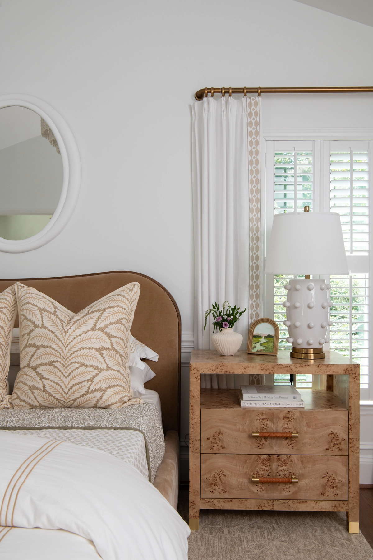 Close-up of the bedside vignette with a burl wood nightstand, white dotted ceramic lamp with brass base, a small vase of flowers, and embroidered sheer curtains beside plantation shutters