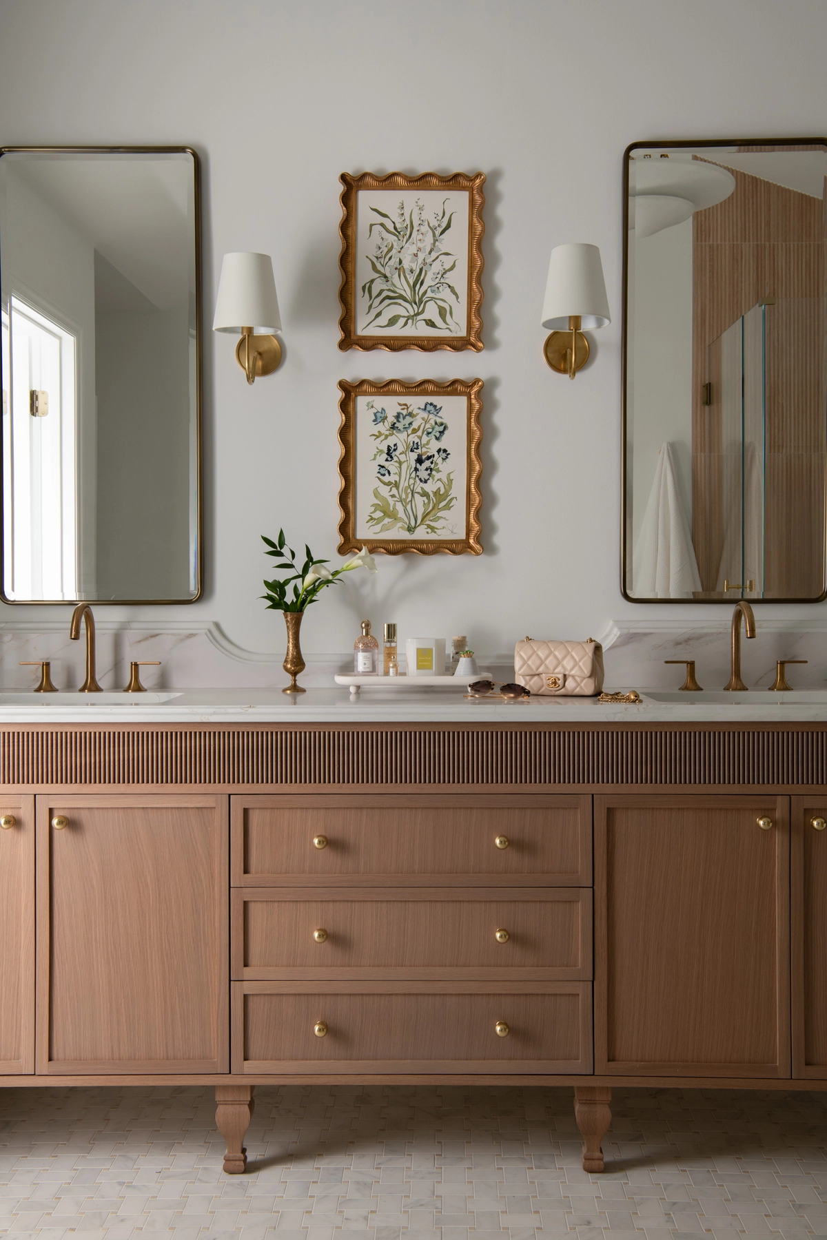 Close-up of the primary bathroom double vanity with warm-toned reeded cabinetry, brass round knobs, white marble countertop, brass faucets, and framed botanical prints in scalloped gold frames