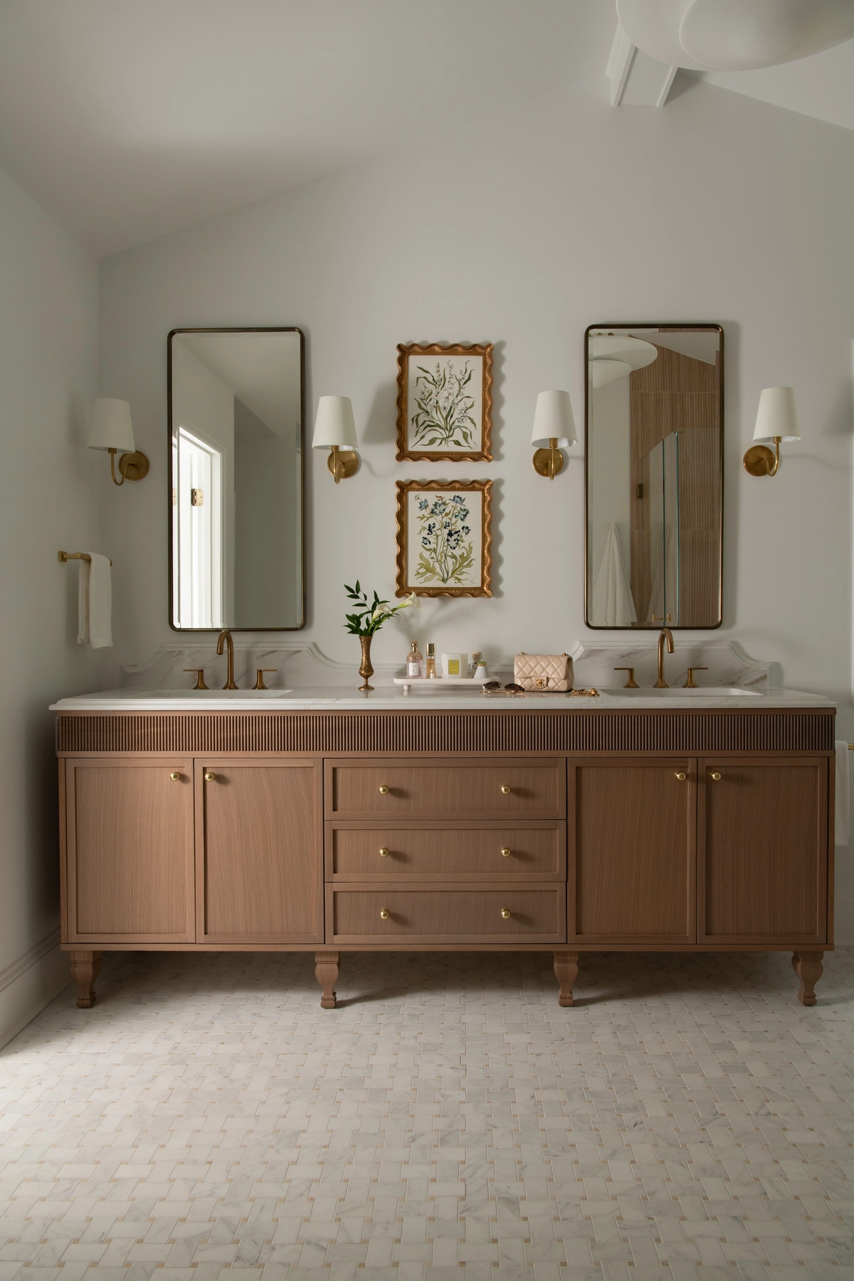 Wide view of the primary bathroom double vanity with warm-toned fluted cabinet, turned legs, brass knobs, white marble countertop, tall rectangular mirrors, and brass sconces