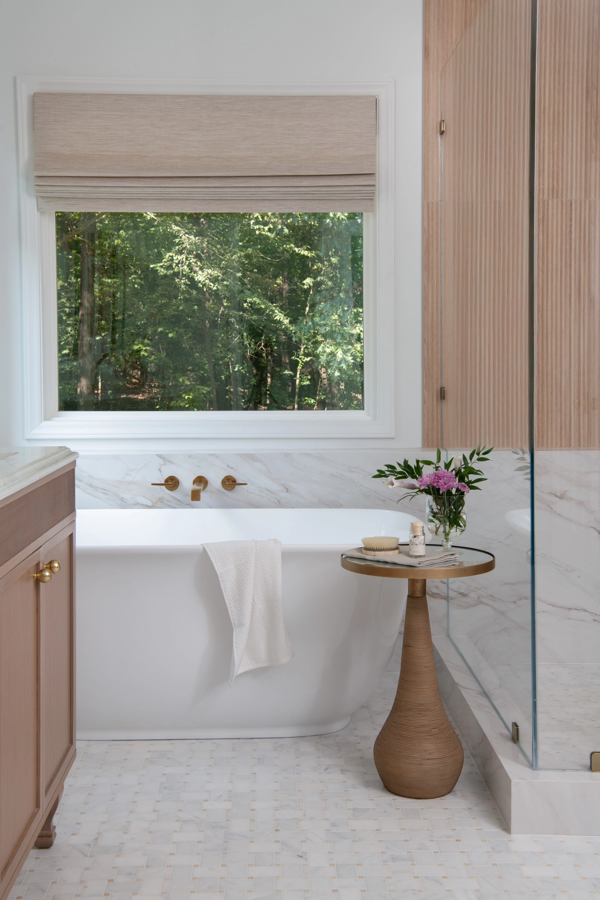 Freestanding white bathtub beneath a large window with a woven Roman shade overlooking lush greenery, flanked by marble wainscoting and a wood pedestal side table with fresh peonies