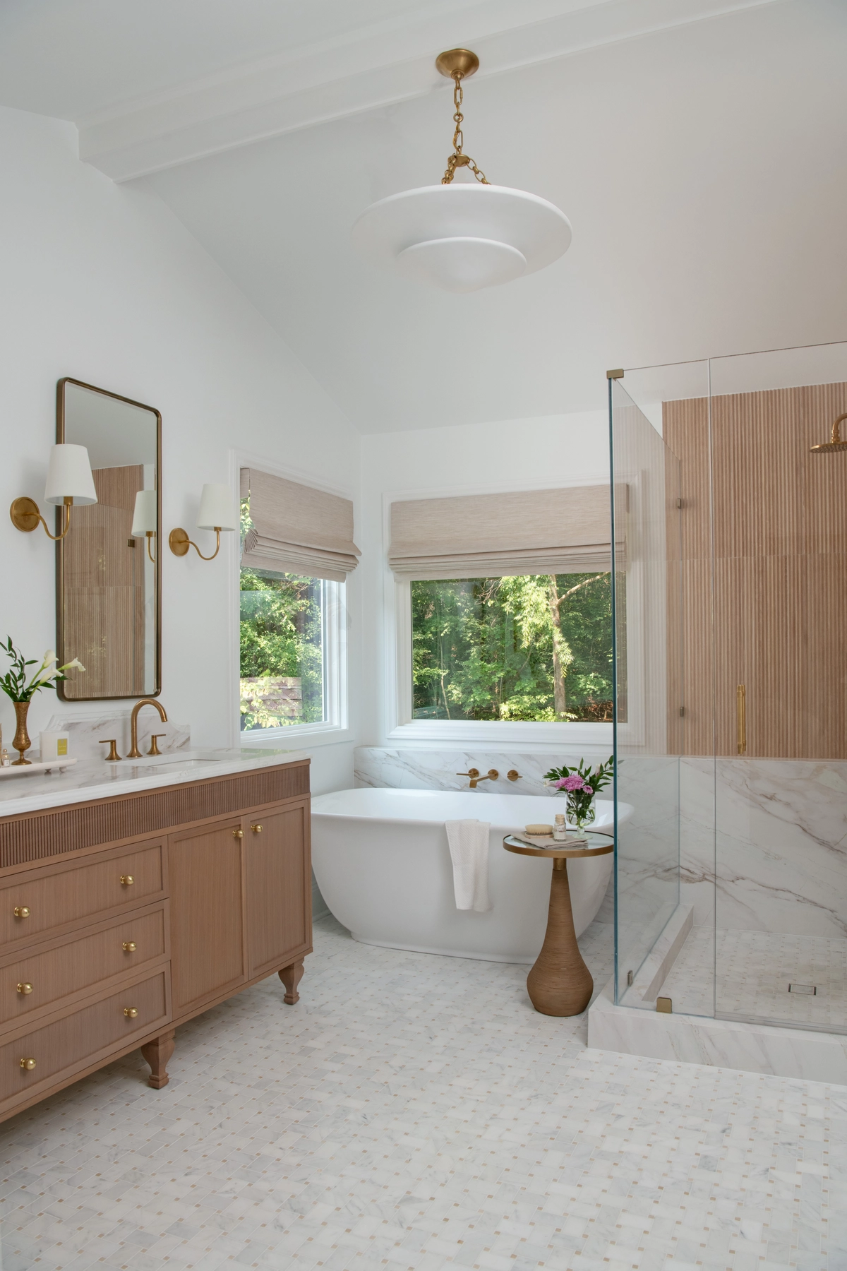 Full view of the guest bathroom with a white pendant light, warm-toned vanity with brass fixtures, a freestanding tub by a large window, and a glass-enclosed shower with fluted wood tile