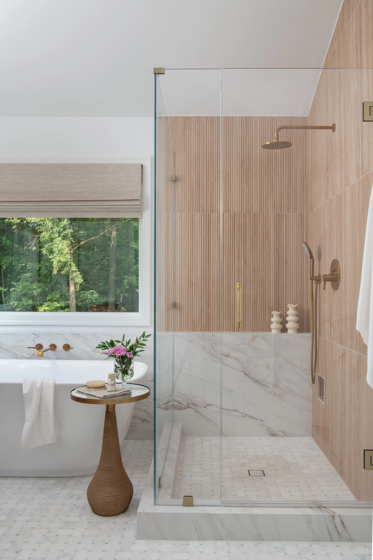 Glass-enclosed shower with fluted light wood tile walls, brass rain showerhead, and marble half-wall, alongside a freestanding white tub beneath a window with greenery
