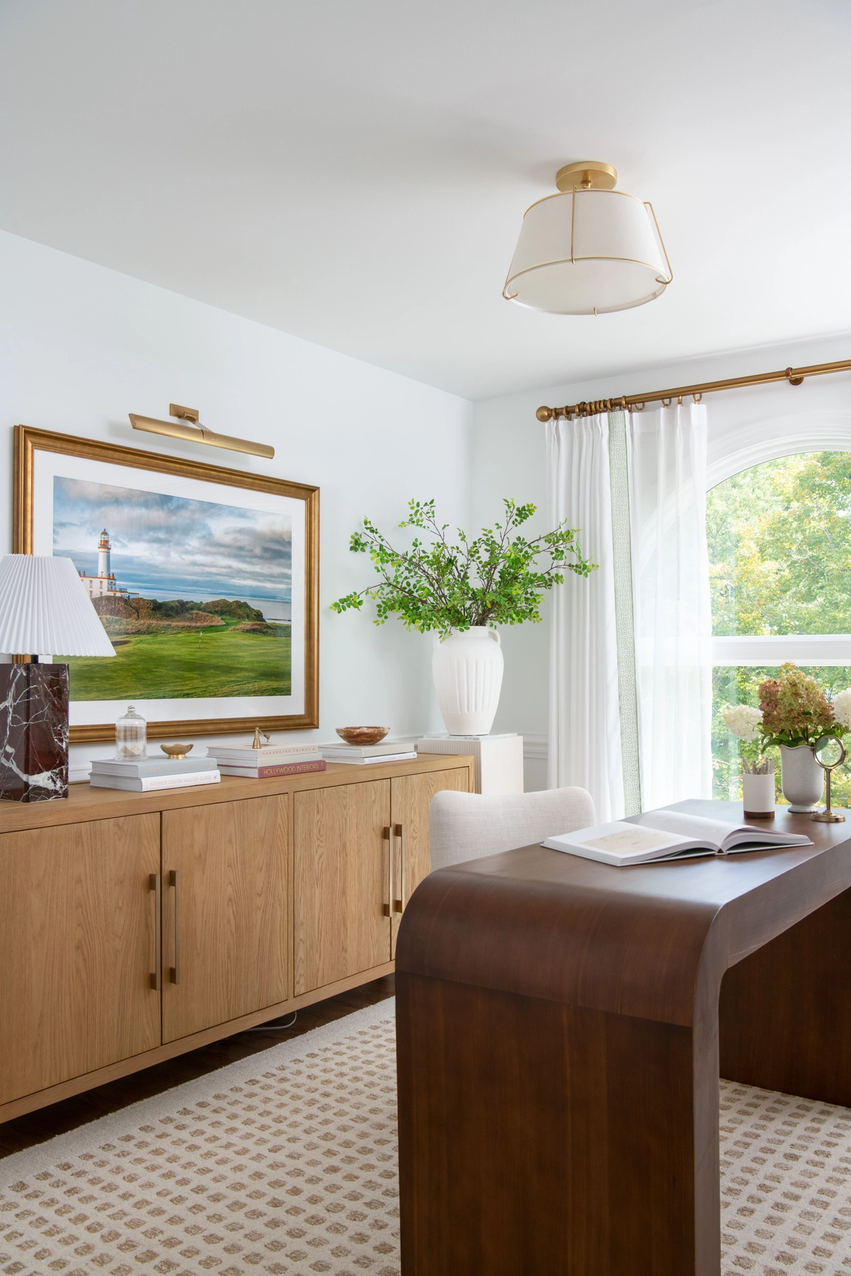 Wide view of the home office showing a light oak credenza beneath a gold-framed lighthouse photograph, a curved walnut waterfall desk, a cream upholstered chair, and a drum pendant light