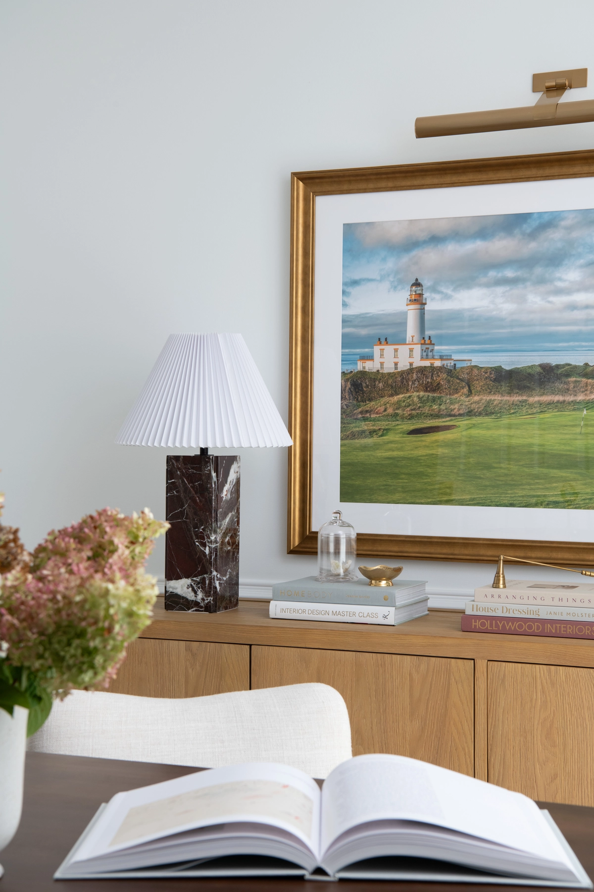 Detail view from the desk looking toward the credenza showing an open book on the walnut surface, a dark marble lamp, a gold-framed lighthouse print, and dried hydrangeas