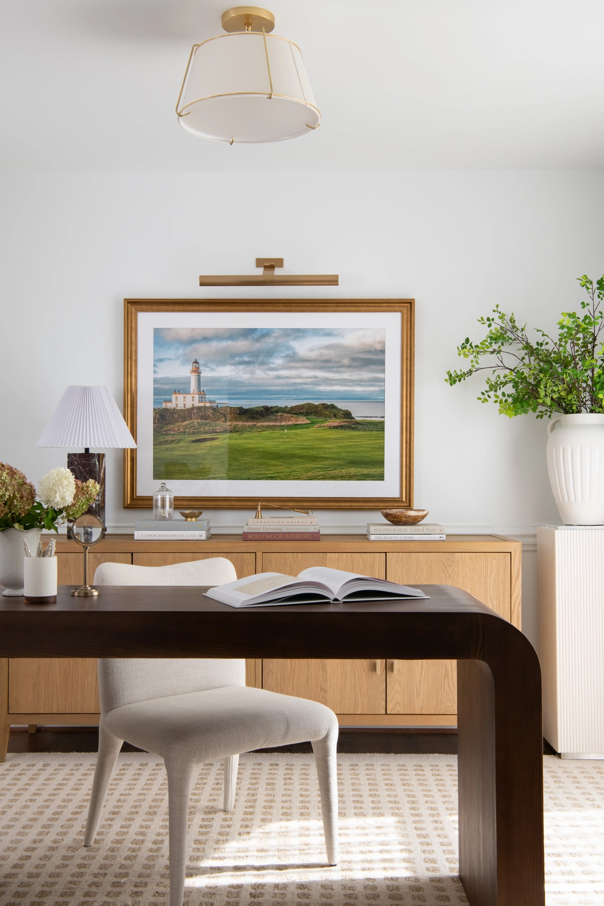 Wide view of the home office centered on the curved walnut waterfall desk with an open book, a cream upholstered chair, and a light oak credenza with a lighthouse print above