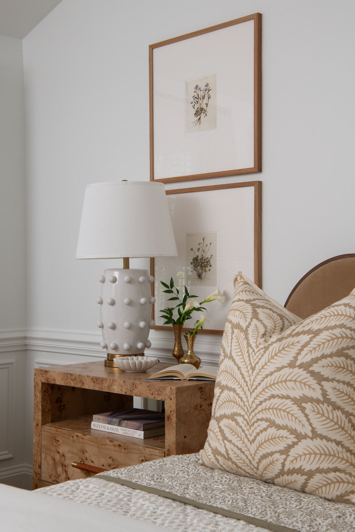 Close-up bedside detail showing a tan botanical leaf pillow against a camel upholstered headboard, a burl wood nightstand with a white dotted ceramic lamp, and framed pressed botanical prints