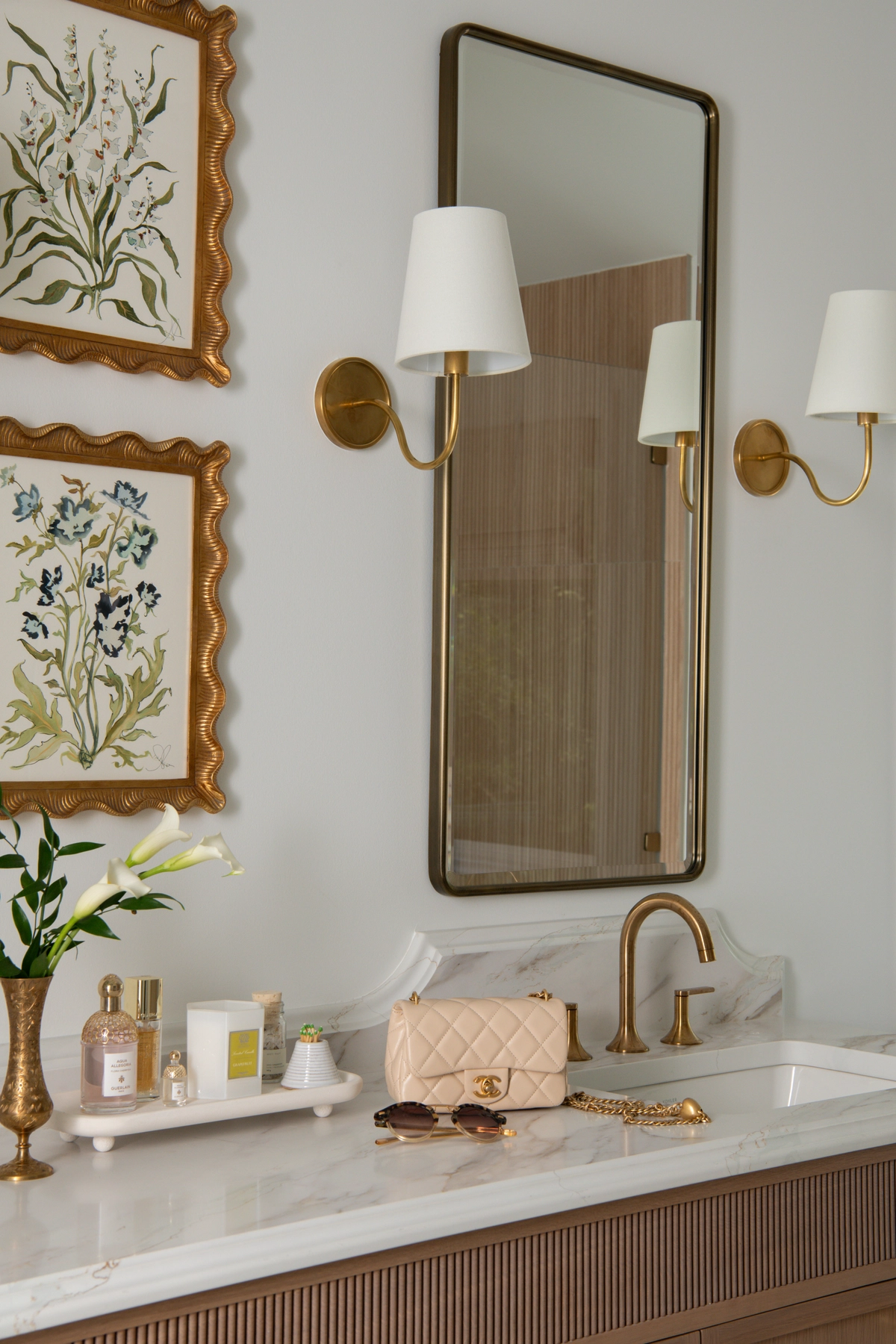 Close-up of the bathroom vanity sink area with a brass gooseneck faucet, white marble countertop, framed botanical print in a scalloped gold frame, and brass wall sconces