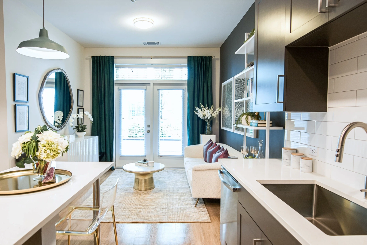 Open-concept model apartment looking from the kitchen island toward the living area, with dark charcoal accent wall, teal curtains, a white sofa with burgundy pillows, and acrylic bar stools