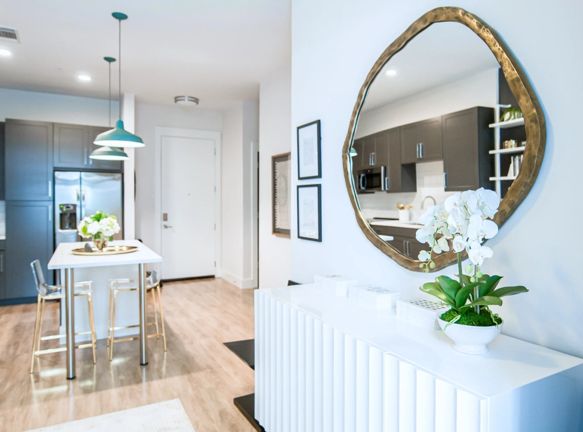 Apartment entryway with a white fluted console table displaying a white orchid, an organic-shaped gold-framed mirror, teal pendant lights over a kitchen island visible in the background
