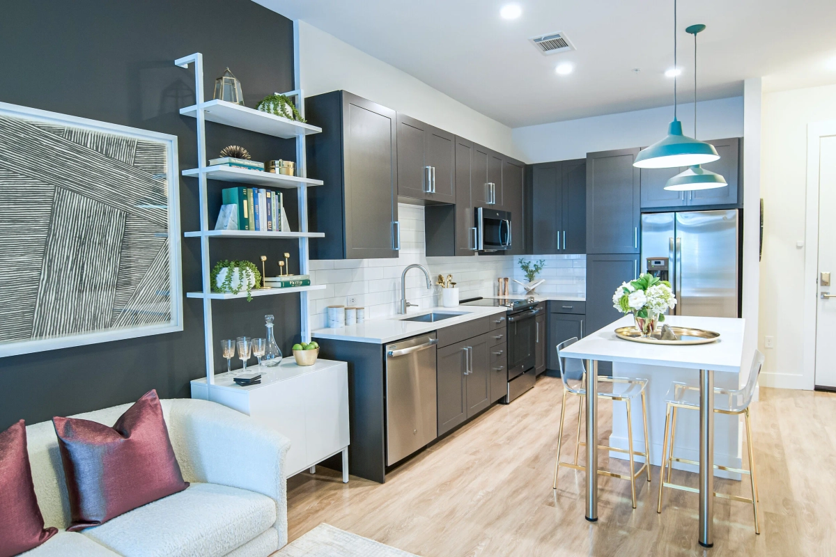 Wide view of the model apartment kitchen featuring dark gray cabinetry, white subway tile backsplash, stainless steel appliances, teal pendant lights, and a small white island with acrylic stools