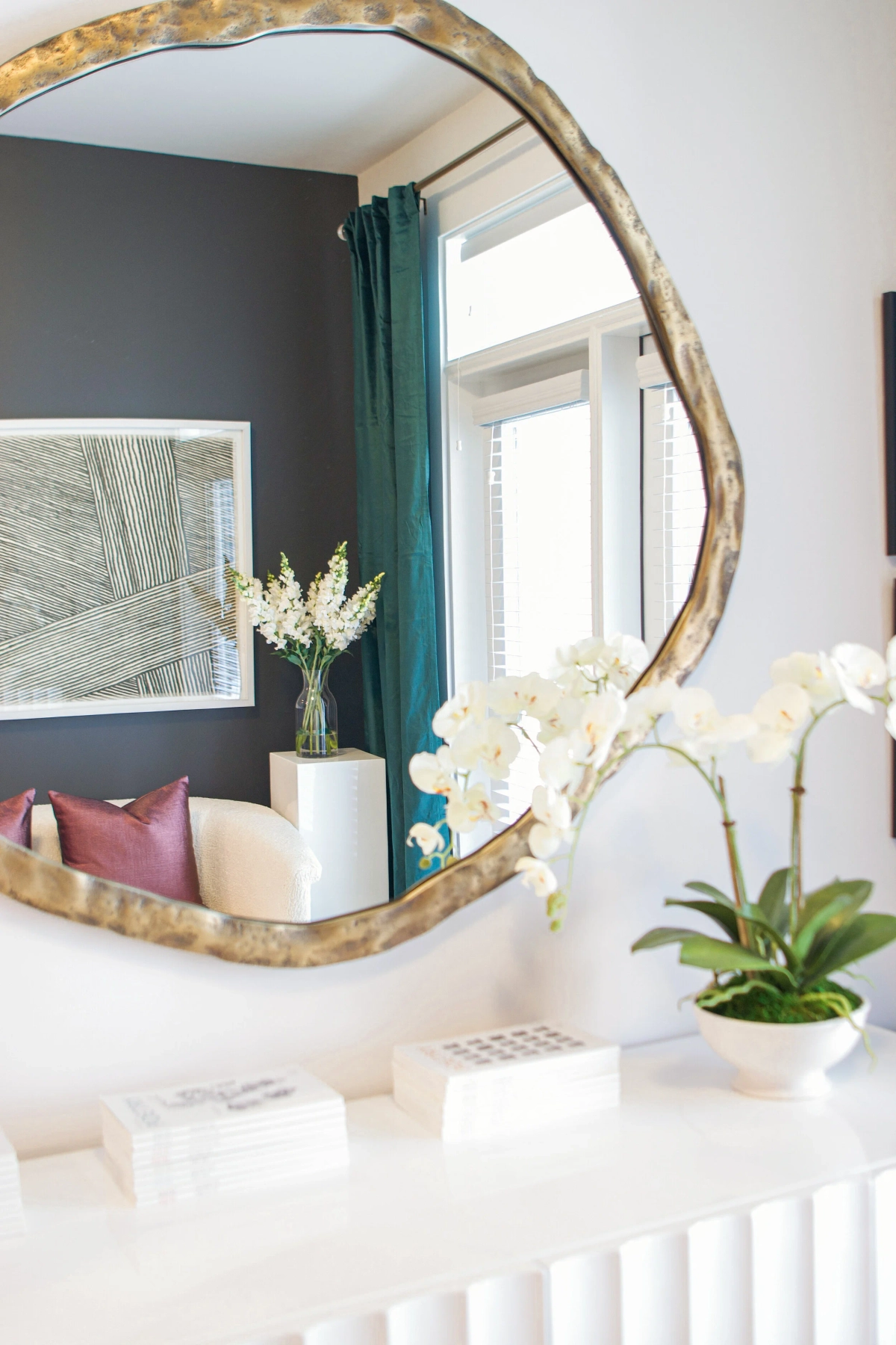 Closeup of an organic-shaped gold-framed mirror reflecting the living room with its dark accent wall and teal curtains, with a white orchid and books on a white fluted console below