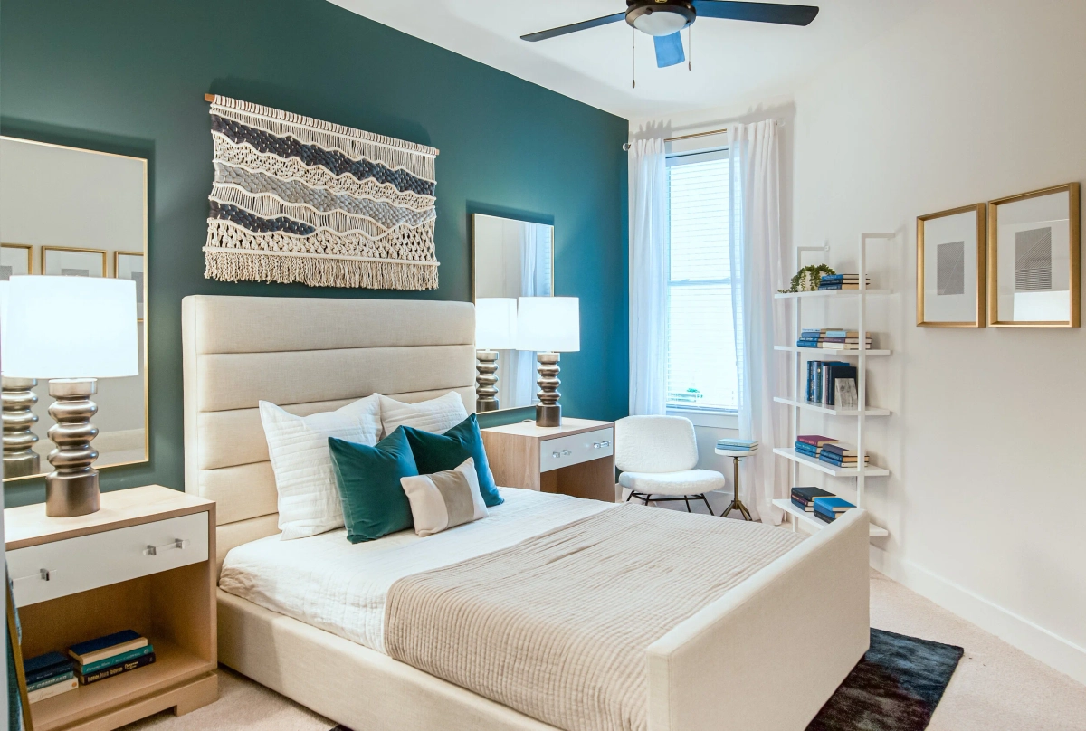 Model bedroom with a teal accent wall, cream upholstered bed with teal and neutral pillows, a woven macrame wall hanging above the headboard, natural wood nightstands, and gold-framed artwork