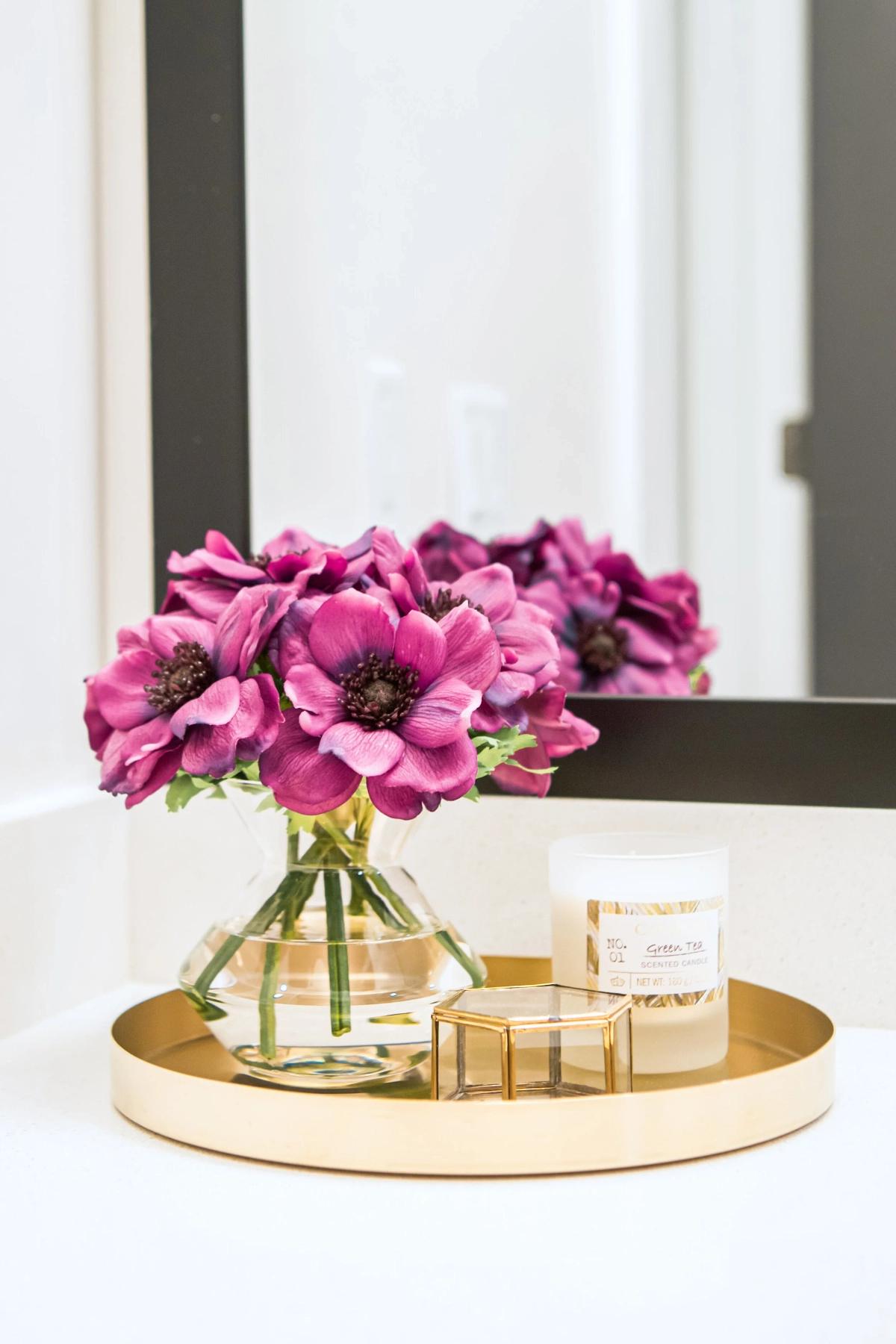 Bathroom vanity detail showing a gold oval tray holding magenta anemone flowers, a cream candle, and a small gold geometric trinket box on a white countertop