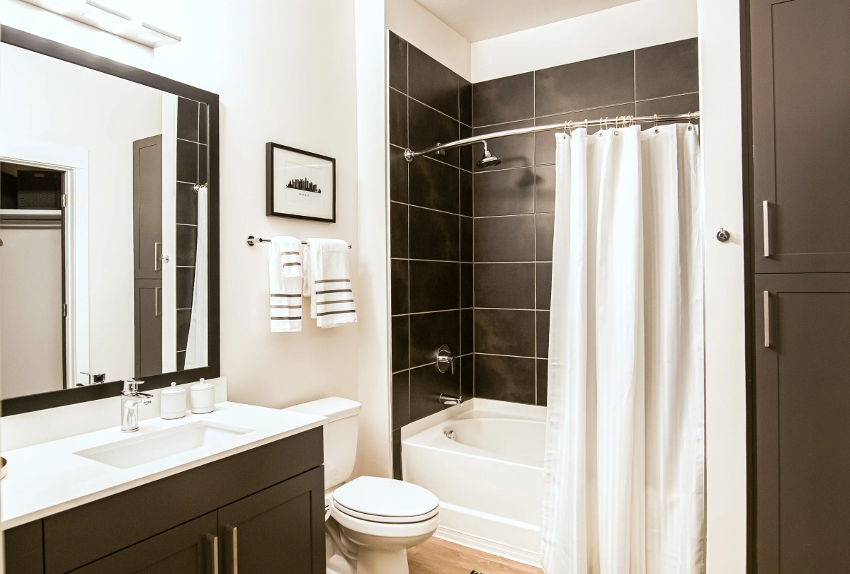 Model apartment bathroom with a dark charcoal tile shower surround, white shower curtain, dark vanity with white countertop, a cityscape art print, and striped towels