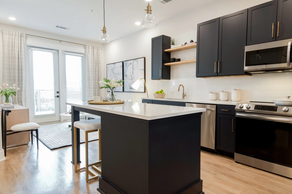 Open-concept kitchen with dark navy cabinetry, white quartz countertop island with gold-legged bar stools, glass pendant lights, stainless steel appliances, and natural wood floating shelves