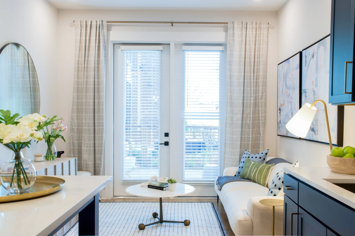 Bright living area with a white sofa accented by blue and green pillows, round marble coffee table, French doors with sheer curtains, and a navy blue credenza with styled accessories