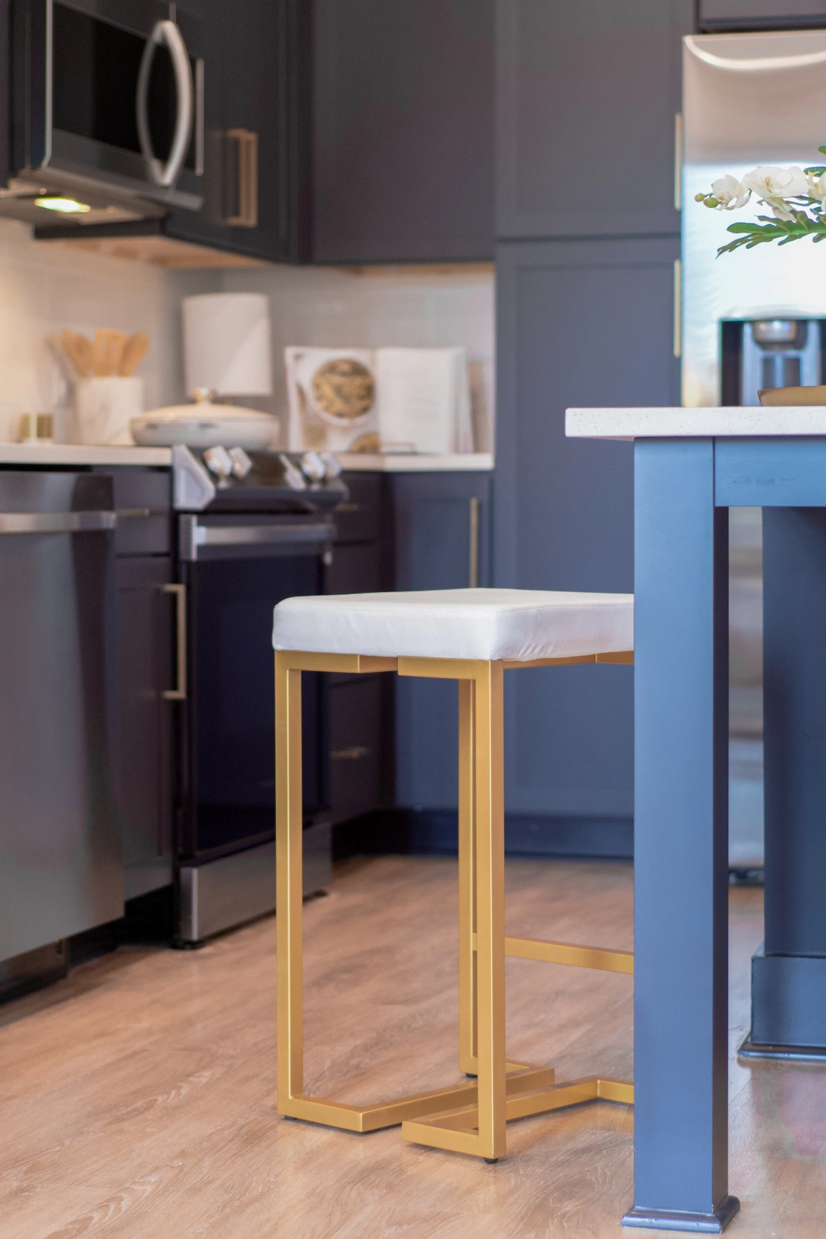 Close-up detail of a white cushioned bar stool with a brass gold base next to a navy blue kitchen island, with dark cabinetry and stainless steel appliances in the background