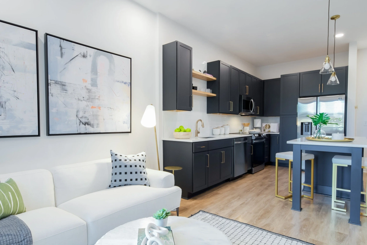 Wide view of an open-concept apartment showing a white sofa with patterned pillows, abstract wall art, and a navy kitchen with floating wood shelves, pendant lighting, and a blue island with gold stools