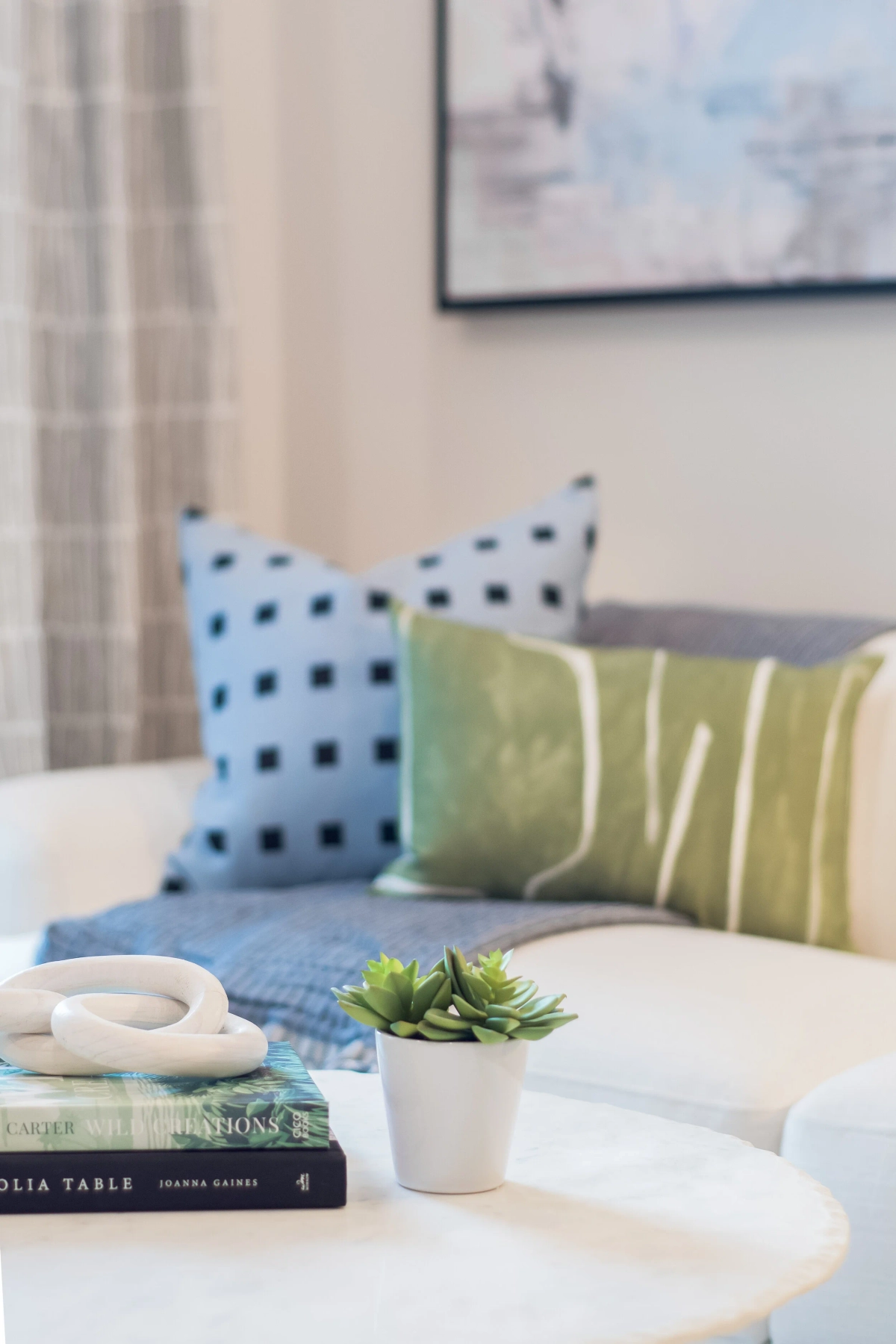 Close-up vignette of a white sofa with blue geometric and green striped accent pillows, a small succulent plant, stacked books, and a decorative chain link object on a round coffee table
