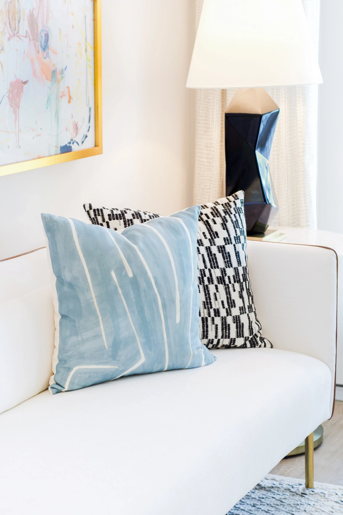 Close-up of a white sofa with a light blue linear-patterned pillow and a geometric pillow, a dark blue sculptural table lamp, and an abstract painting in the background