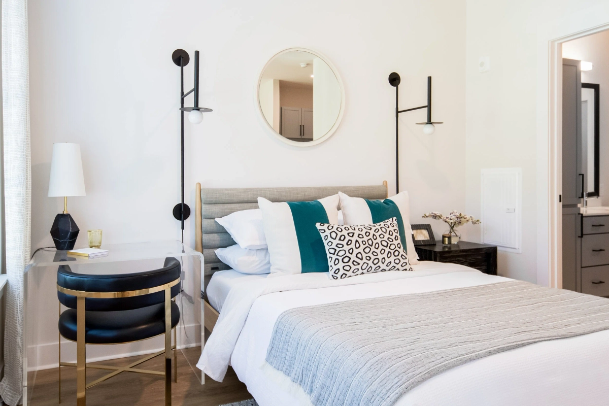 Studio apartment bedroom with a gray upholstered headboard, teal and leopard-print accent pillows, a round mirror, black modern wall sconces, a navy leather accent chair with brass frame