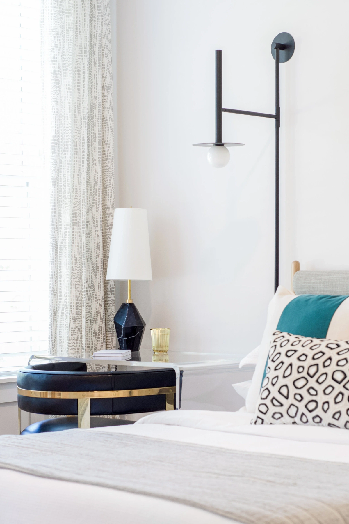 Bedroom nightstand detail showing a black modern wall sconce, a dark blue faceted table lamp, a navy leather and brass accent chair, teal and patterned pillows on white bedding