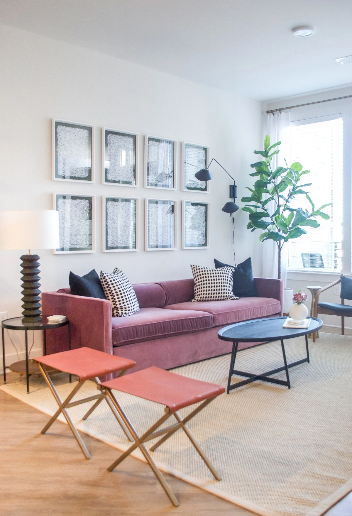 Living room with a mauve velvet sofa, black-and-white patterned throw pillows, a grid of six framed abstract prints, coral leather folding stools, a black oval coffee table, and a fiddle leaf fig tree