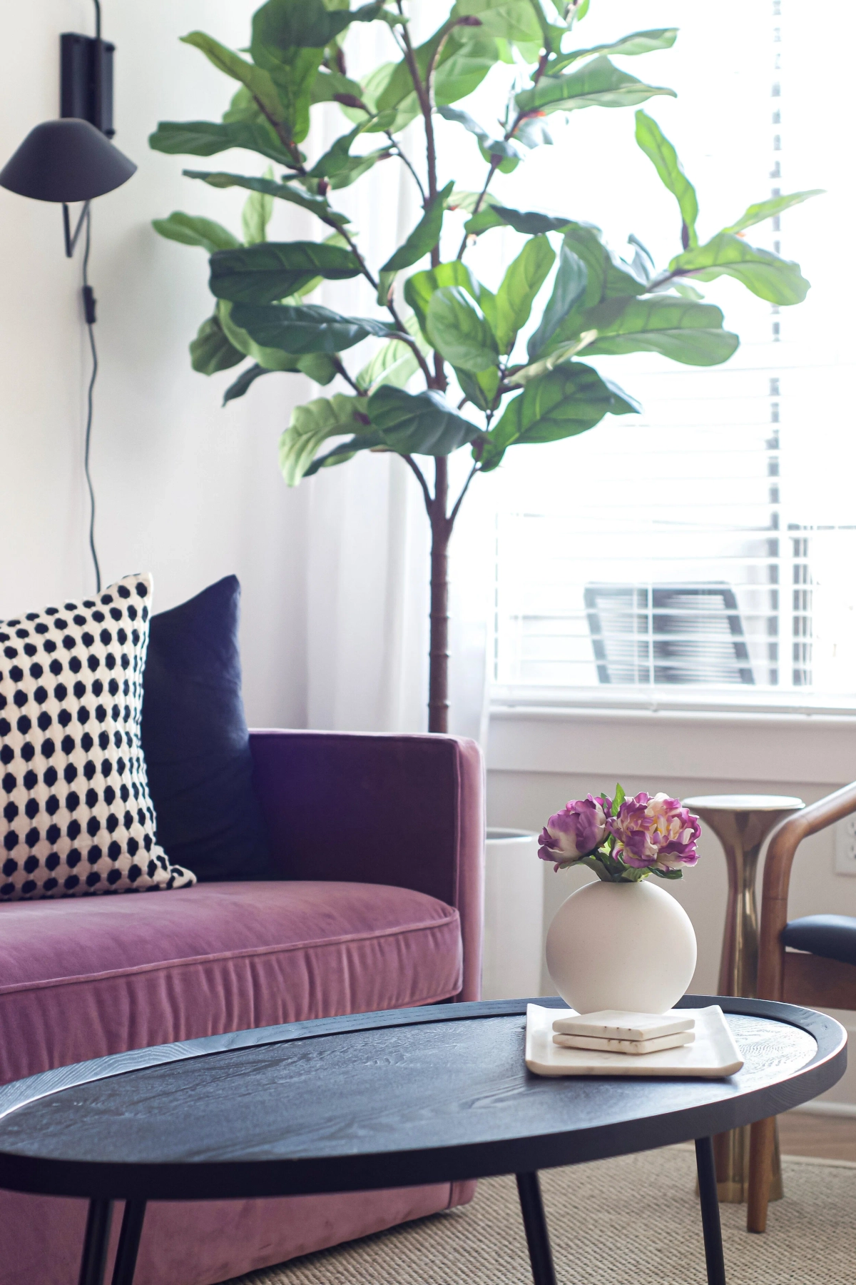 Close-up of a mauve velvet sofa corner with a polka dot pillow, a black oval coffee table with a ceramic vase of pink flowers, and a fiddle leaf fig tree by the window