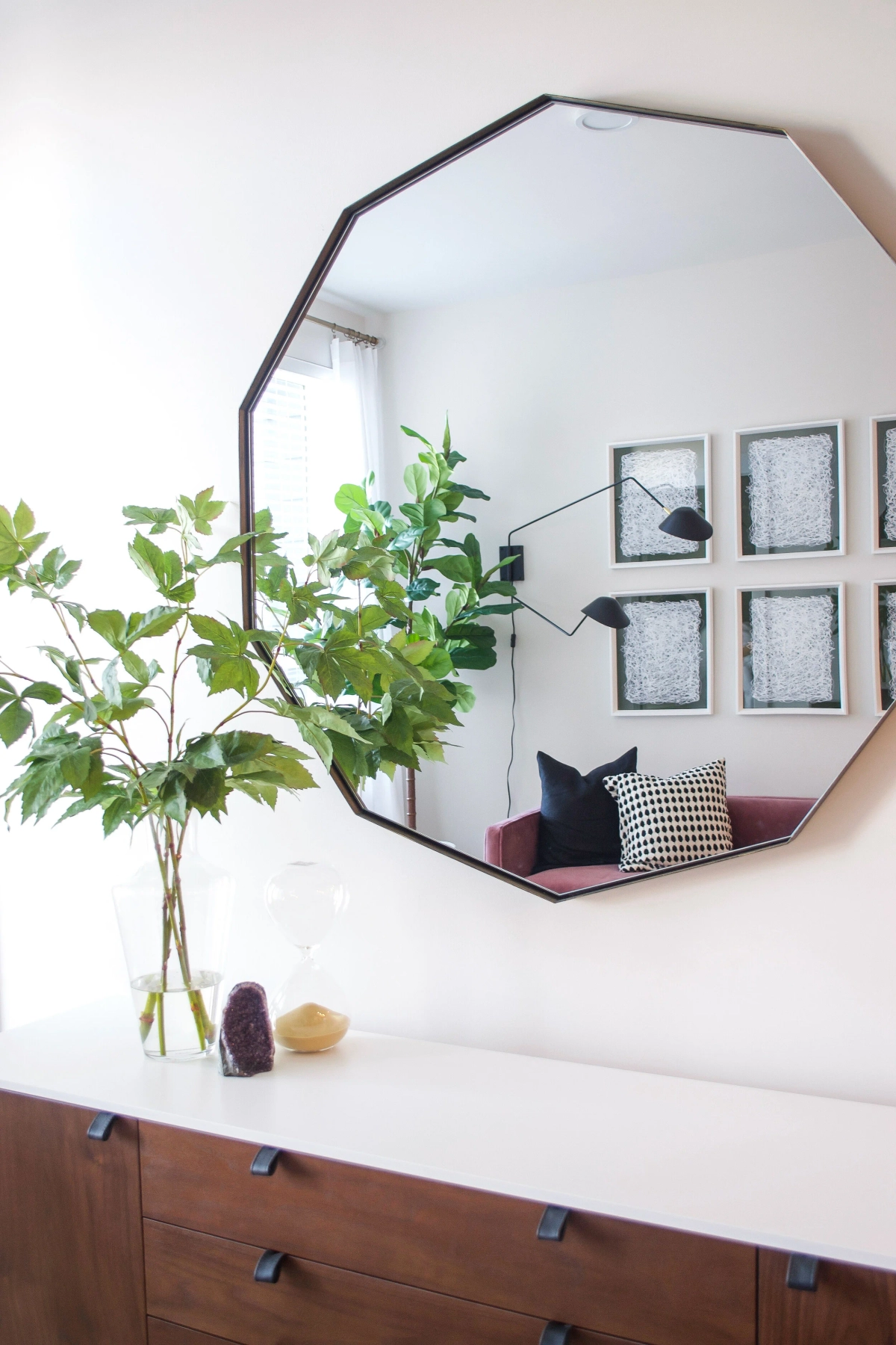 Close-up of a large geometric octagonal mirror on the wall above a mid-century walnut credenza, reflecting the living room with its mauve sofa and gallery wall