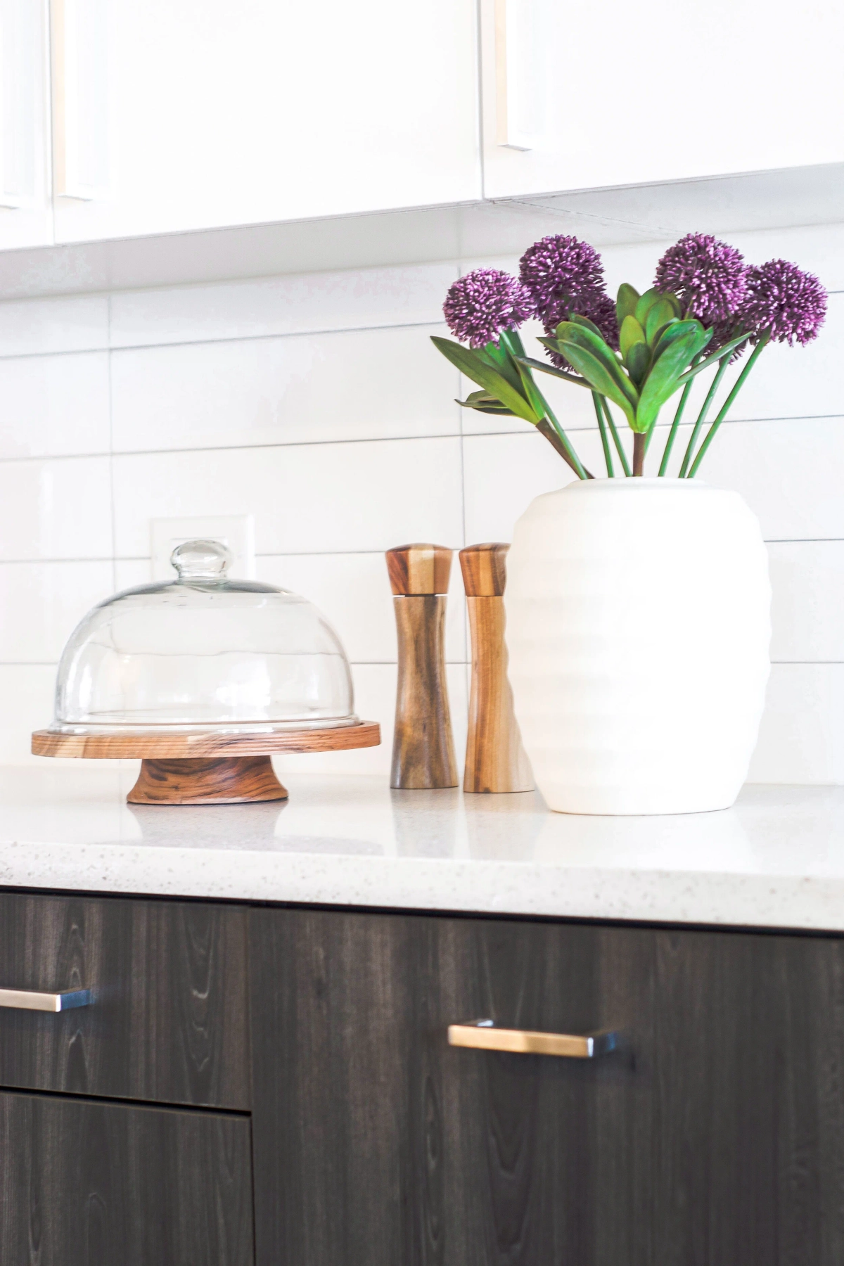 Kitchen counter detail with dark wood cabinetry, brass hardware, a white quartz countertop, a glass cake dome, wooden salt and pepper mills, and a white vase with purple allium flowers