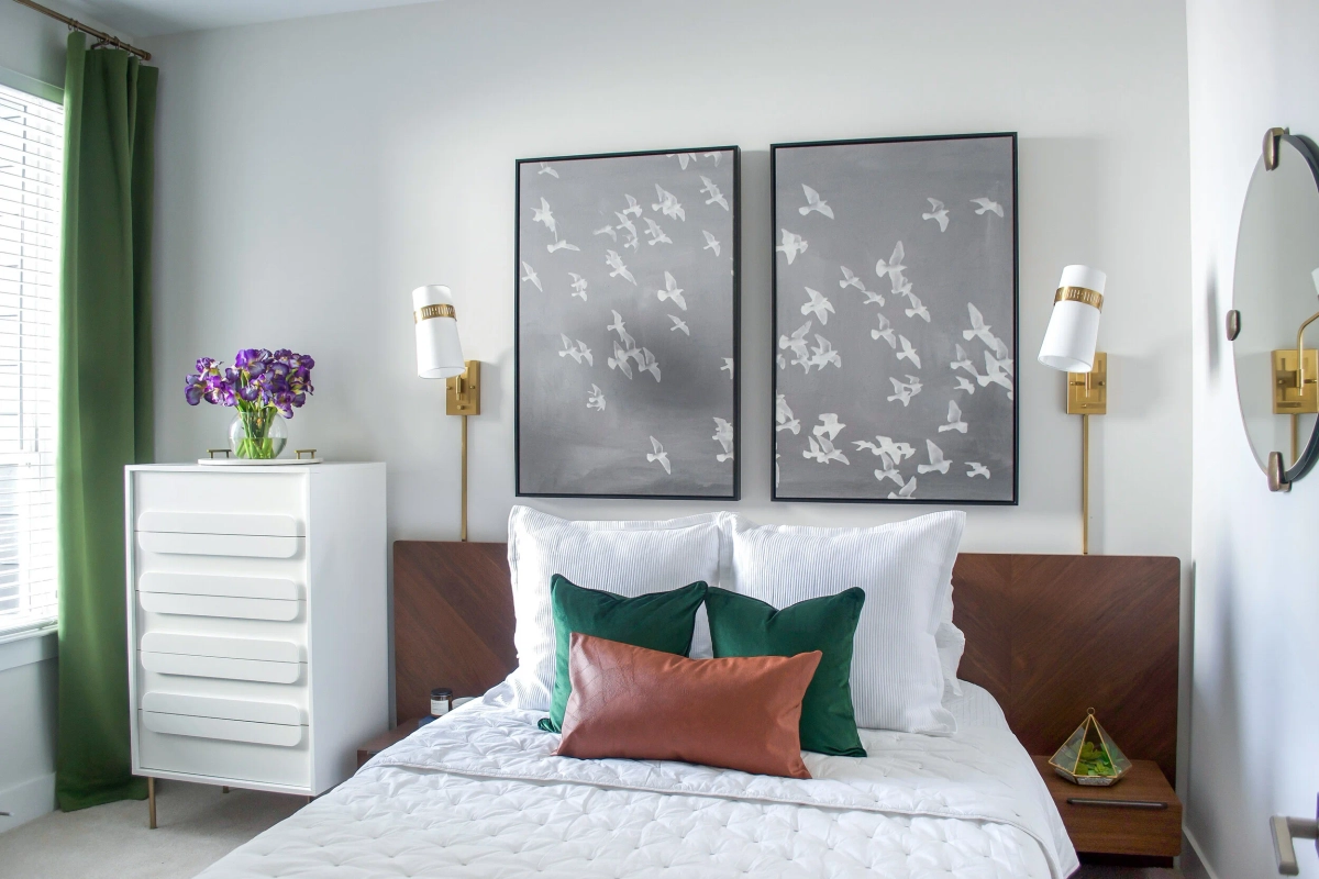 Bedroom with a walnut chevron headboard, white quilted bedding, emerald green and rust accent pillows, brass wall sconces, gray bird-motif artwork, green curtains, and a white dresser