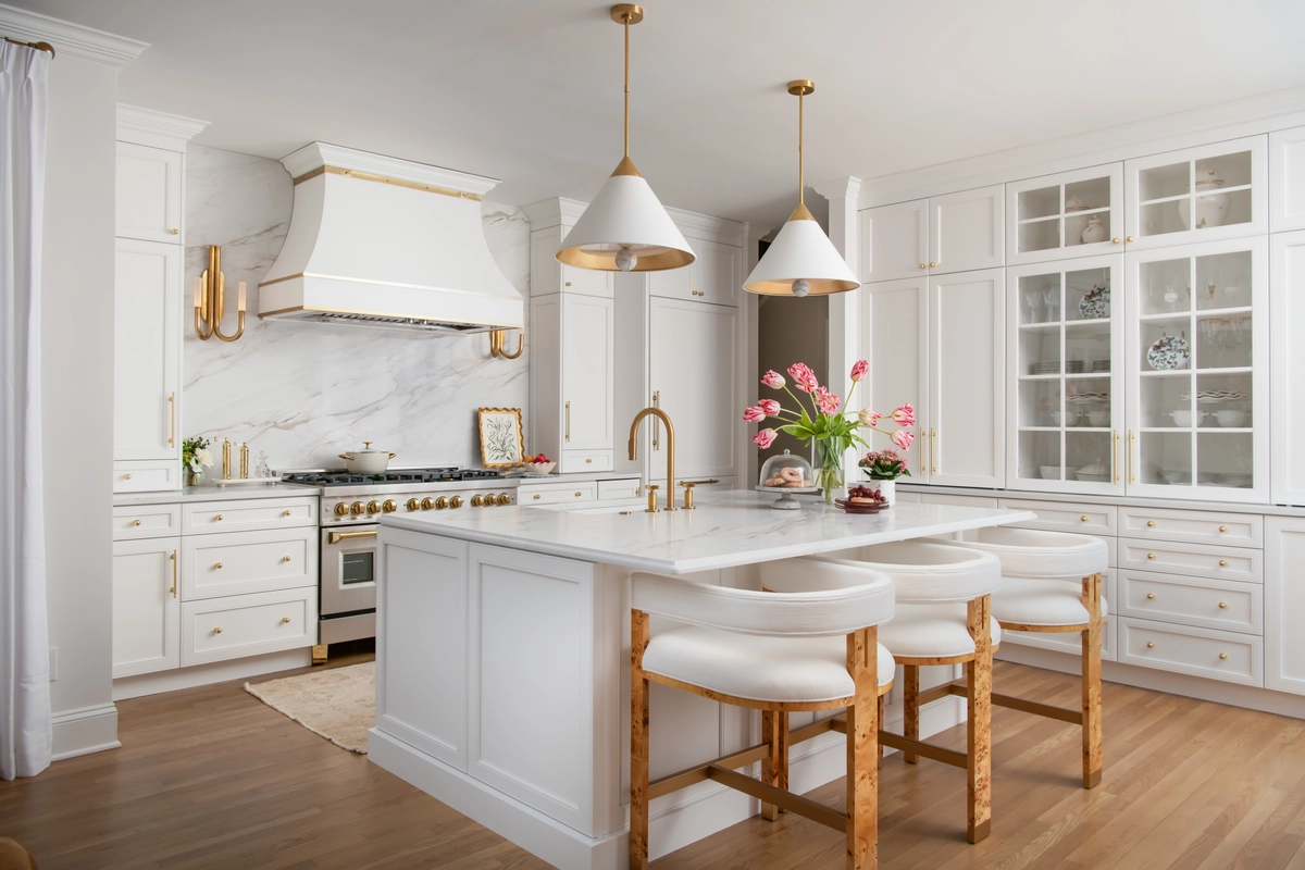 White kitchen island with marble countertop, burl wood bar stools, brass pendants, and glass-front cabinetry